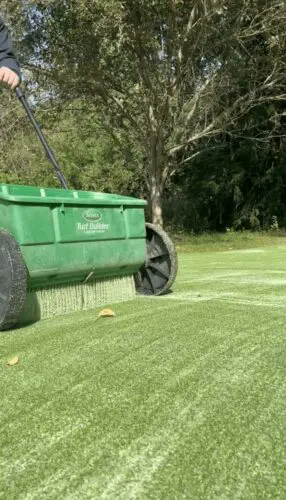 Low-angle photograph of a technician applying fine, cooling infill sand to a newly installed artificial grass lawn.