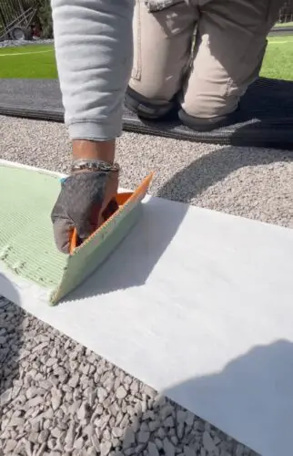 A close-up of a professional artificial turf installation showing a technician applying specialized green adhesive onto industrial seam tape between two sections of synthetic grass.