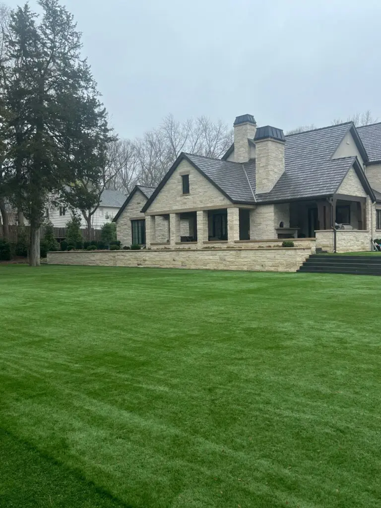 A large, perfectly manicured green artificial turf lawn installed in front of a modern stone estate with stone retainers and dark patio steps.